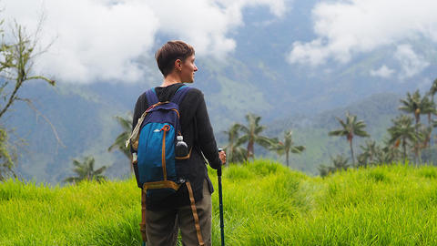 View of woman standing, holding 2 hiking sticks, with blue backpack on. Face in profile as looking at mountains and palm trees