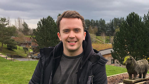 Jordan Johnson, sitting on boulder in front of gray sky, field, trees, and "Brompton Lakes" sign