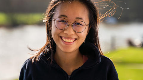 Photo of Kaylin Chong, wearing glasses outdoors on breezy day, blurred background
