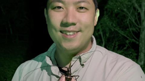 Sang smiling in woods at night, holding monochamus odaesan beetle toward camera, green light from below